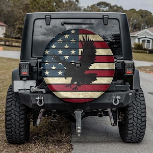 May include: A black Jeep with a spare tire cover featuring the American flag design. The cover displays the flag's stars and stripes, with a silhouette of an eagle in the center. The Jeep is parked on a paved road.