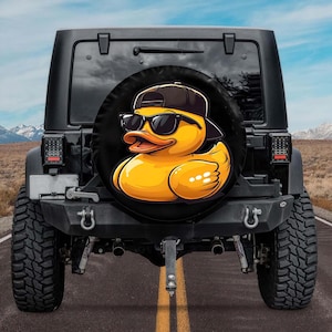 May include: Black spare tire cover featuring a cartoon yellow duck wearing sunglasses and a black cap. The duck has an orange beak and is set against a black background. The cover is mounted on the back of a black Jeep.