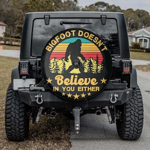 May include: Black Jeep with a spare tire cover featuring a Bigfoot silhouette against a sunset design. The cover includes the text "Bigfoot Doesn't Believe In You Either" in a circular layout, with stars.