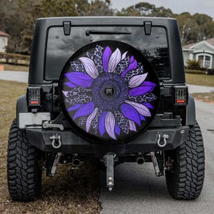 May include: Black Jeep with a spare tire cover featuring a large purple sunflower design. The sunflower has purple petals and a leopard print pattern. The vehicle is parked on a paved road.