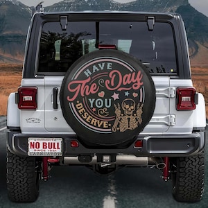 May include: A black spare tire cover with the text "Have The Day You Deserve" in pink and white, along with a skeleton graphic. The cover is on a white Jeep with a license plate that reads "NO BULL".