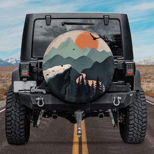 May include: A black Jeep with a spare tire cover featuring a mountain landscape design. The cover displays a sunset, mountains, and trees in shades of orange, green, and gray. The Jeep is parked on a paved road with a desert backdrop.