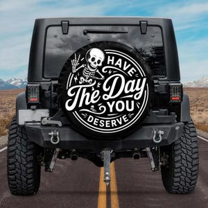 May include: Black spare tire cover with a white skeleton graphic giving a peace sign. The cover has the text "Have The Day You Deserve" in a circular design. The cover is mounted on the back of a black Jeep.
