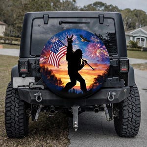 May include: Black Jeep with a spare tire cover featuring a silhouette of Bigfoot holding an American flag and a peace sign. The background includes fireworks and a sunset scene. The Jeep has large tires.