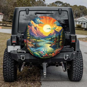 May include: A black Jeep with a colorful spare tire cover featuring a landscape scene. The cover displays a river winding through mountains under a bright sun and clouds. The Jeep has large tires.