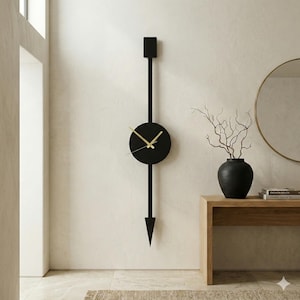Puede incluir: Un reloj de pared negro moderno con manecillas doradas. El reloj tiene una esfera circular y un diseño vertical largo, con una parte superior cuadrada y una inferior puntiaguda. Está montado en una pared de color claro.