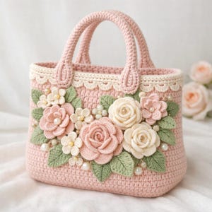 Pode incluir: Uma mala de mão rosa de crochê com duas alças. A mala é decorada com flores de crochê em tons de rosa e creme, folhas verdes e pequenos detalhes de pérolas. A borda superior apresenta uma borda de renda creme.