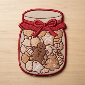 May include: An embroidered patch shaped like a glass jar filled with various holiday-themed cookies, including a gingerbread person, a Christmas tree, and snowflake shapes. The jar features a red fabric lid cover tied with a matching red bow.