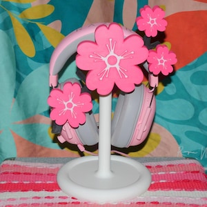 May include: A pink and gray headset rests on a white headphone stand adorned with pink flower decorations. The stand is on a pink and white striped surface, with a colorful patterned background.