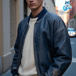 Könnte beinhalten: Blaue Leder-Bomberjacke mit geripptem Kragen und Bündchen. Die Jacke hat einen Reißverschluss vorne und einen cremefarbenen Pullover darunter. Goldene Initialen sind auf dem linken Ärmel gestickt. Die Jacke wird im Freien getragen.