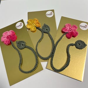 Peut inclure: Trois marque-pages faits à la main en forme de fleurs au crochet. Chaque marque-page présente une fleur colorée en rose, jaune ou rose, avec une tige et une feuille vertes. Les marque-pages sont présentés sur des cartes dorées.