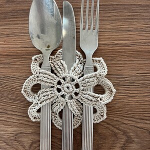 May include: A vintage cutlery set, comprising a spoon, knife, and fork, rests on a beige crocheted doily. The cutlery features a ridged handle design. The background is a wooden surface.
