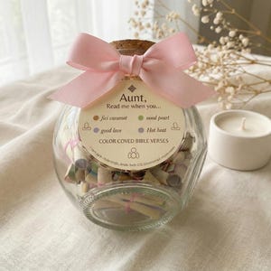 Puede incluir: Un tarro de cristal transparente lleno de papeles enrollados, atados con gomas rosas. El tarro tiene un tapón de corcho y un lazo rosa. Una etiqueta dice "Aunt, Read me when you..." con frases codificadas por colores. Una pequeña vela blanca está cerca.