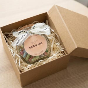 Może przedstawiać: Szklany słoik wypełniony kolorowymi przedmiotami, zawiązany białą wstążką i umieszczony w brązowym pudełku prezentowym. Słoik ma niestandardową etykietę z tekstem i małą ilustracją. Pudełko jest otwarte, ukazując zawartość.