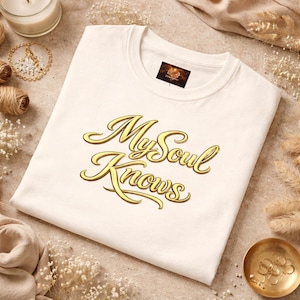 Może przedstawiać: Kremowa koszulka z napisem "My Soul Knows" w złotym skrypcie. Koszulka ma okrągły dekolt i małą metkę przy szyi. Koszulka jest złożona i umieszczona na neutralnej powierzchni.