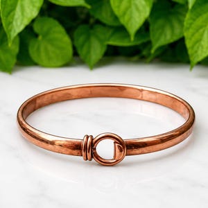 May include: A polished copper bangle bracelet featuring a smooth, rounded band and a circular hook-and-eye style clasp mechanism. The metal has a warm, reddish-brown metallic finish with a reflective surface, forming a continuous circular shape.