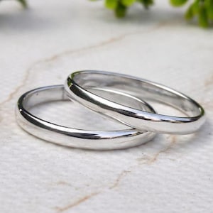 Può includere: Due fedi nuziali d'argento, una sull'altra. Gli anelli sono lisci e lucenti, riflettendo la luce. Sono appoggiati su una superficie di marmo bianco e beige, con un fogliame verde sullo sfondo.