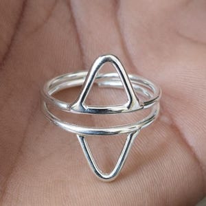 Könnte beinhalten: Ein silberner Ring mit geometrischem Design. Der Ring hat zwei Dreiecke, eines zeigt nach oben und das andere nach unten, verbunden durch ein Band. Der Ring besteht aus glänzendem Silbermetall.