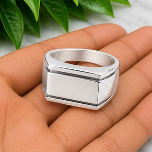 Peut inclure: Une bague chevalière en métal argenté poli avec une surface supérieure plate et rectangulaire ainsi qu'un anneau large et solide. La bague présente une finition métallique propre avec des bords nets et définis, offrant un design simple et structurel.