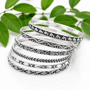 May include: A stack of silver bangles with intricate floral and geometric patterns. The bangles are arranged in a slightly curved stack, showcasing the detailed designs. Green leaves provide a natural backdrop.