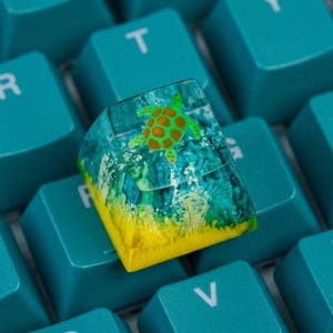 Pode incluir: Uma tecla personalizada de resina com uma seção superior transparente contendo uma figura de tartaruga marinha verde e laranja, sobre uma base azul e amarela que imita uma cena subaquática, posicionada em um teclado verde-azulado.