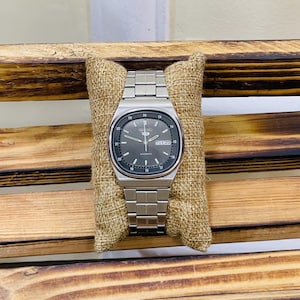 Puede incluir: Un reloj de pulsera automático Seiko plateado con una esfera rectangular y una correa de metal a juego. La esfera del reloj es de color gris oscuro con marcadores de hora y agujas blancas. El día y la fecha se muestran a las 3 en punto.