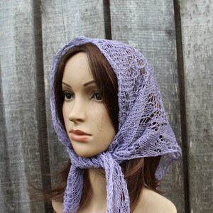 May include: A lilac-coloured, open-weave headscarf is draped over a mannequin's head and tied at the neck. The scarf has a delicate, lacy pattern and is made of a soft, lightweight material. The background is a weathered wooden fence.