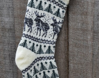 Könnte beinhalten: Cremefarbener gestrickter Weihnachtsstrumpf mit einem Muster aus dunkelgrünen Bäumen und dunkelgrauen Elchen. Der Bund und die Spitze sind einfarbig cremefarben. Der Strumpf hängt vor einem verwitterten Holzgrund.