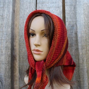 May include: A knitted hood in shades of red, orange, and brown. The hood is worn by a mannequin and is made of a textured yarn. The hood is designed to cover the head and neck, with a scarf-like extension that can be tied or draped.