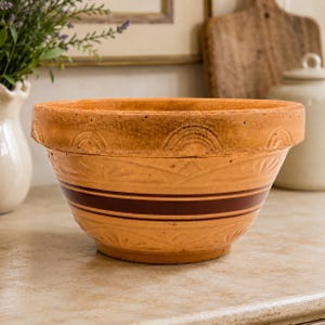 May include: A vintage-style, tan ceramic mixing bowl with two dark brown stripes. The bowl has a wide rim with embossed arch designs and a detailed floral pattern on the sides. This farmhouse-style bowl is perfect for kitchen decor.