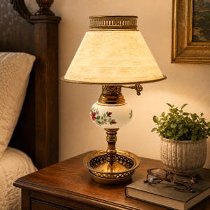 May include: A vintage-style bedside lamp with a cream-colored lampshade and a brass base. The lamp features a white globe with red and green floral detailing. It sits on a wooden nightstand next to a small potted plant and a pair of eyeglasses.