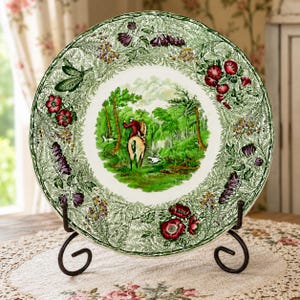 May include: A round ceramic plate featuring a central illustration of a person on horseback riding through a green forest with dogs. The wide border is decorated with a detailed botanical pattern of red flowers, purple blossoms, and green foliage.