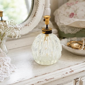 May include: A clear glass perfume bottle with a rounded, vertically ribbed body and a metallic gold-toned pump mechanism. The bottle features a textured surface and a dark, weathered-looking collar connecting the glass base to the gold top.