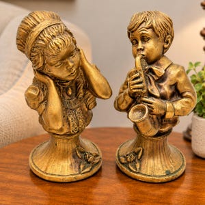 May include: Two antique gold-colored statues. One depicts a girl with her hands on her ears, the other a boy playing a saxophone. Both statues have detailed clothing and are mounted on decorative bases with floral accents. The statues are likely made of a metal alloy.