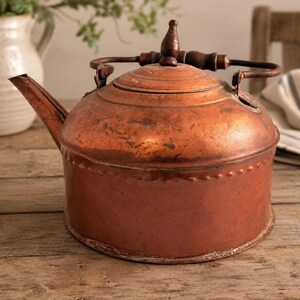 May include: A vintage copper kettle with a wooden handle and lid finial. The kettle has a rounded body, a spout, and a handle. The copper has a warm, reddish-brown color, and the kettle appears to be aged. The kettle is likely used for boiling water.