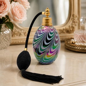May include: An oval glass perfume bottle featuring a swirling iridescent pattern of green, purple, and white colors. The bottle is topped with a gold-toned metal atomizer cap and includes a black fabric bulb pump with a matching black tassel.