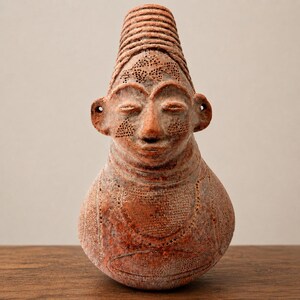 May include: A terracotta-colored ceramic sculpture shaped like a human head and torso. The piece features a tall, ribbed conical headdress, prominent ears with holes, and intricate stippled patterns across the face and chest area.