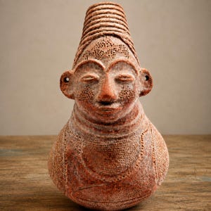 May include: A terracotta-colored ceramic vessel shaped like a human head with a tall, ribbed conical headdress. The surface features intricate stippled textures, etched patterns, and small circular ear details, with a weathered, matte finish.