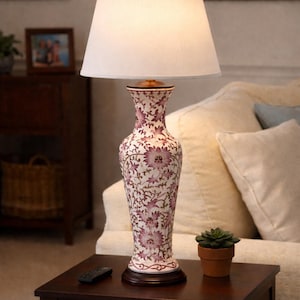 May include: A decorative table lamp with a white lampshade and a floral patterned base. The base is a tall, vase-like shape with a cream background and pink floral designs. The lamp sits on a dark brown wooden table.