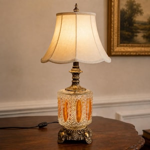May include: A table lamp with a cream-colored fabric shade and a brass-toned metal base. The lamp features a square glass body with amber-colored oval insets and a textured pattern. The base is decorated with floral motifs and a black power cord.