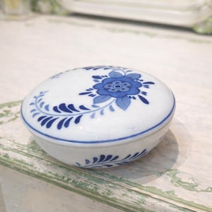 May include: A white ceramic trinket box with a blue floral design on the lid. The box has a round shape and is decorated with blue floral and leaf patterns. The box is likely used for storing small items.