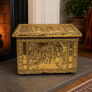 May include: A rectangular brass chest featuring intricate embossed scenes of figures in a village setting on the front panel and lid. The metal surface has a polished gold tone with detailed decorative borders framing the central relief imagery.