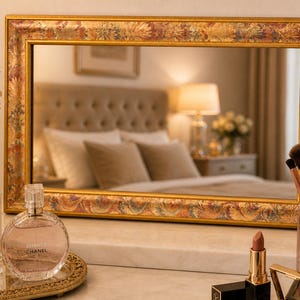 May include: A rectangular gold framed mirror with floral and leaf designs. The mirror reflects a bedroom scene with a bed, lamp, and pillows. Perfume bottles, makeup brushes, and lipstick are on the vanity.