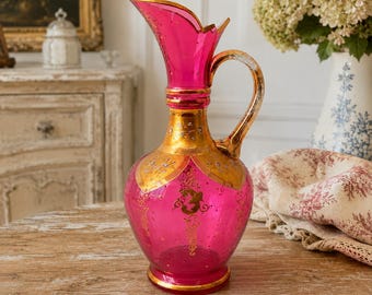 May include: A vibrant magenta glass pitcher featuring an ornate gold-painted neck and handle. The body is decorated with intricate gold filigree and floral patterns, with a wide, flared rim and a curved handle extending from the neck to the shoulder.