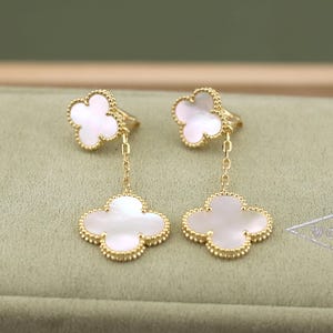 May include: Gold-tone clover-shaped earrings with white mother-of-pearl inlays. The earrings feature a delicate chain connecting the clover to the ear post. The clover design is outlined with a beaded border, adding a touch of elegance.