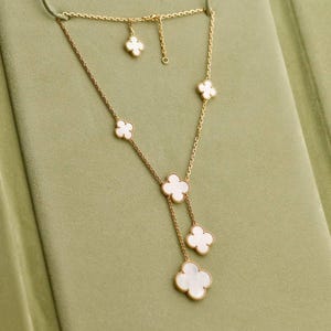 以下が含まれることがあります： 白いクローバー型のペンダントが付いたゴールドカラーのネックレス。 ネックレスは、さまざまなサイズの複数のクローバーチャームを備えた繊細なチェーンデザインで、レイヤード効果を生み出しています。 ネックレスは、柔らかな緑色の背景に表示されています。