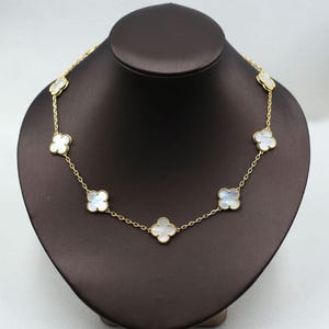 May include: A gold-colored necklace featuring a series of white clover-shaped pendants. The necklace is displayed on a brown jewelry bust. The chain is delicate, and the pendants have a pearlescent sheen.