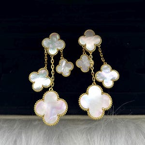 May include: Dangling earrings featuring four-leaf clover designs. Each earring has multiple clover-shaped pendants in white mother-of-pearl, outlined in gold. The pendants are connected by delicate gold chains, creating a layered effect.