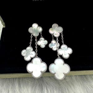May include: Elegant silver-tone drop earrings featuring four-leaf clover designs. Each earring has four clover-shaped pendants with a mother-of-pearl finish, connected by delicate chains. The earrings are set against a black background.