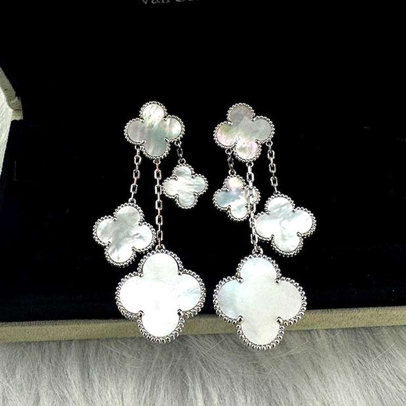 May include: Elegant silver-tone drop earrings featuring four-leaf clover designs. Each earring has four clover-shaped pendants with a mother-of-pearl finish, connected by delicate chains. The earrings are set against a black background.